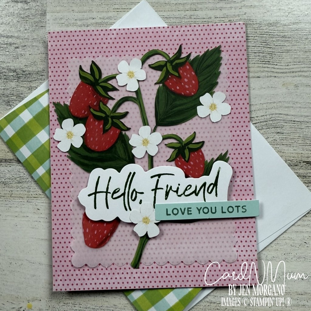 A greeting card featuring strawberries and flowers with the text 'Hello, Friend' and 'Love You Lots' on a pink dotted background. The card is placed on a wooden surface alongside a green checkered envelope.