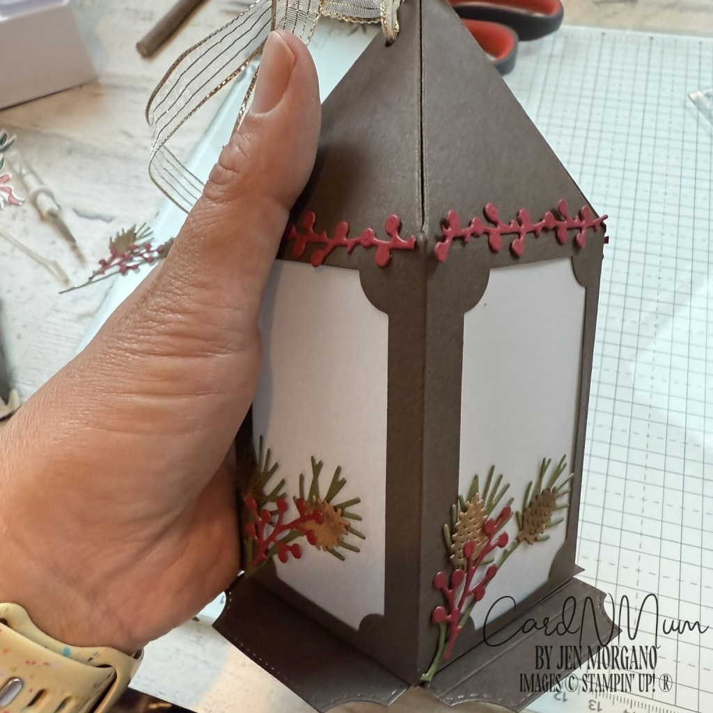 A person holding a decorative lantern made of cardstock, featuring festive embellishments and a ribbon at the top.