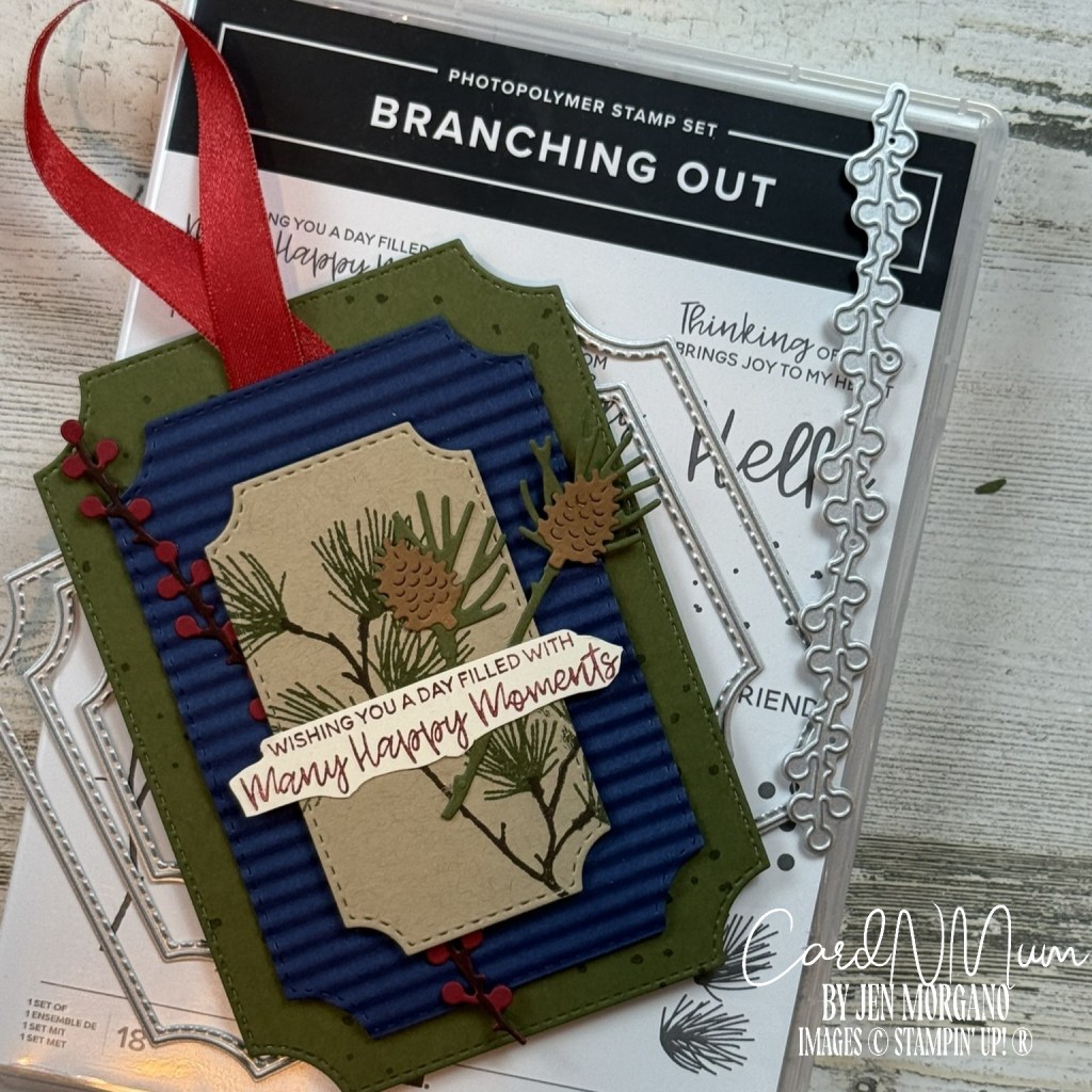 A handcrafted tag featuring a stitched design with a layered die-cut in green and blue, adorned with pinecones and branches, and a red ribbon, placed on top of a 'Branching Out' stamp set packaging.