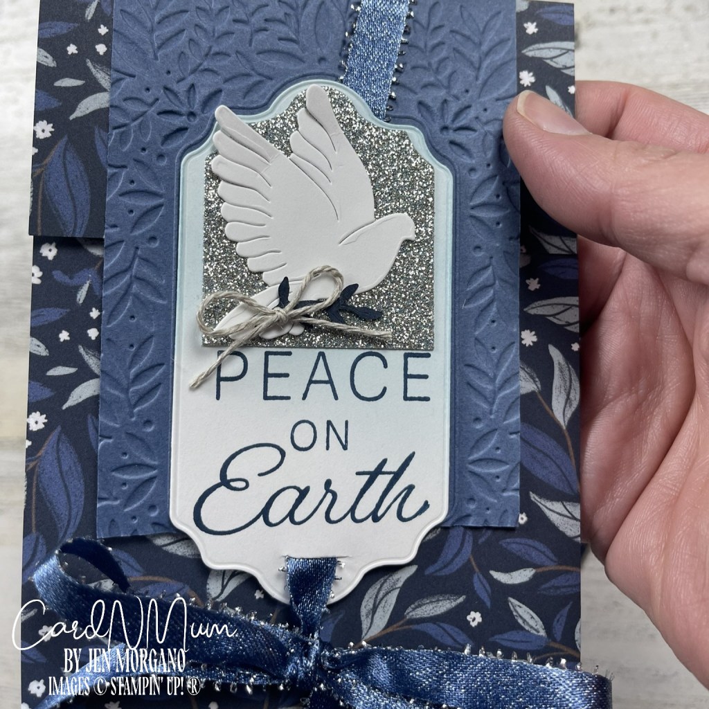 A handmade greeting card featuring a white dove on a glittery background, with the message 'PEACE ON EARTH' in blue letters, adorned with a decorative ribbon and floral patterned paper.