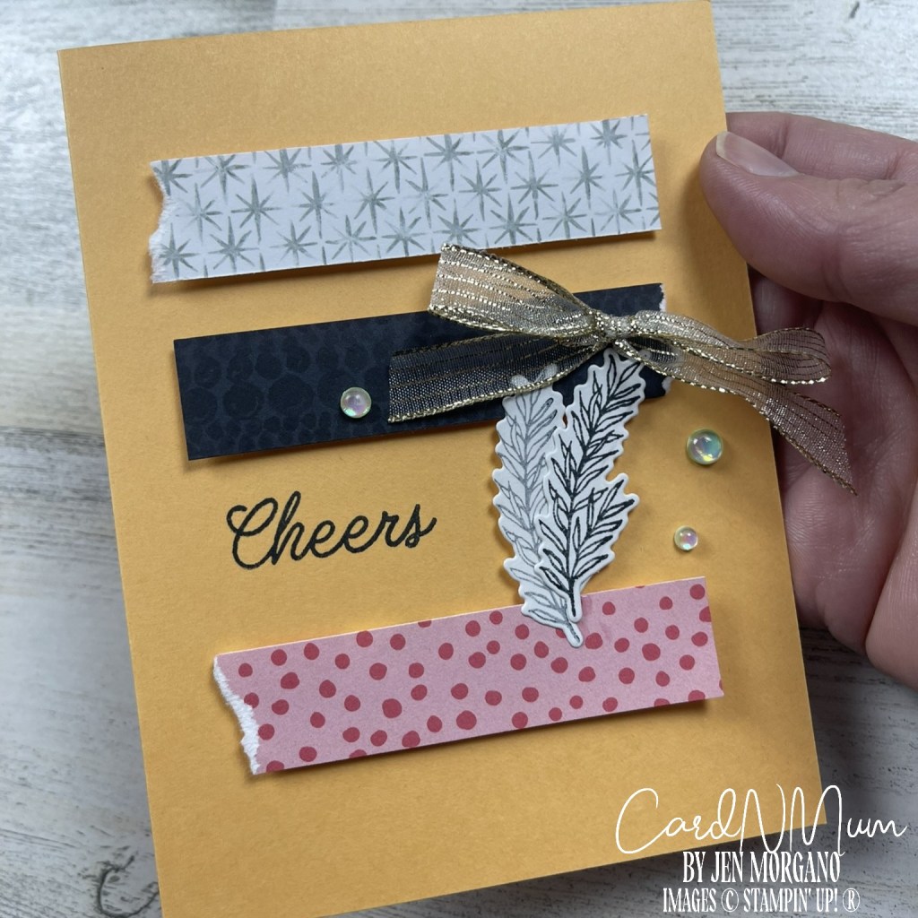 Close-up of a handmade card featuring a yellow background with the word 'Cheers' in black. The card includes various decorative strips in patterns, a silver ribbon bow, and a leafy element.