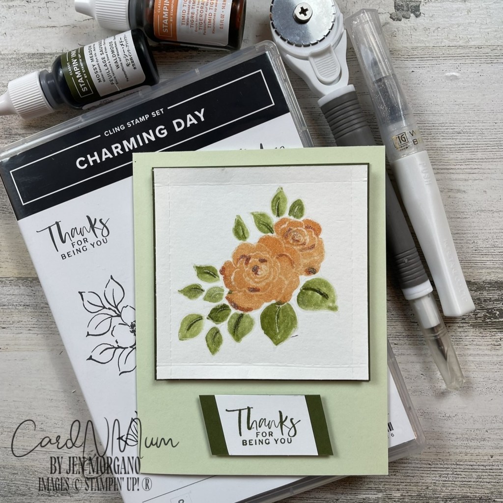 A handmade greeting card featuring orange roses and green leaves, with the message 'Thanks for being you' displayed prominently. The card is paired with various crafting supplies, including ink pads and markers.