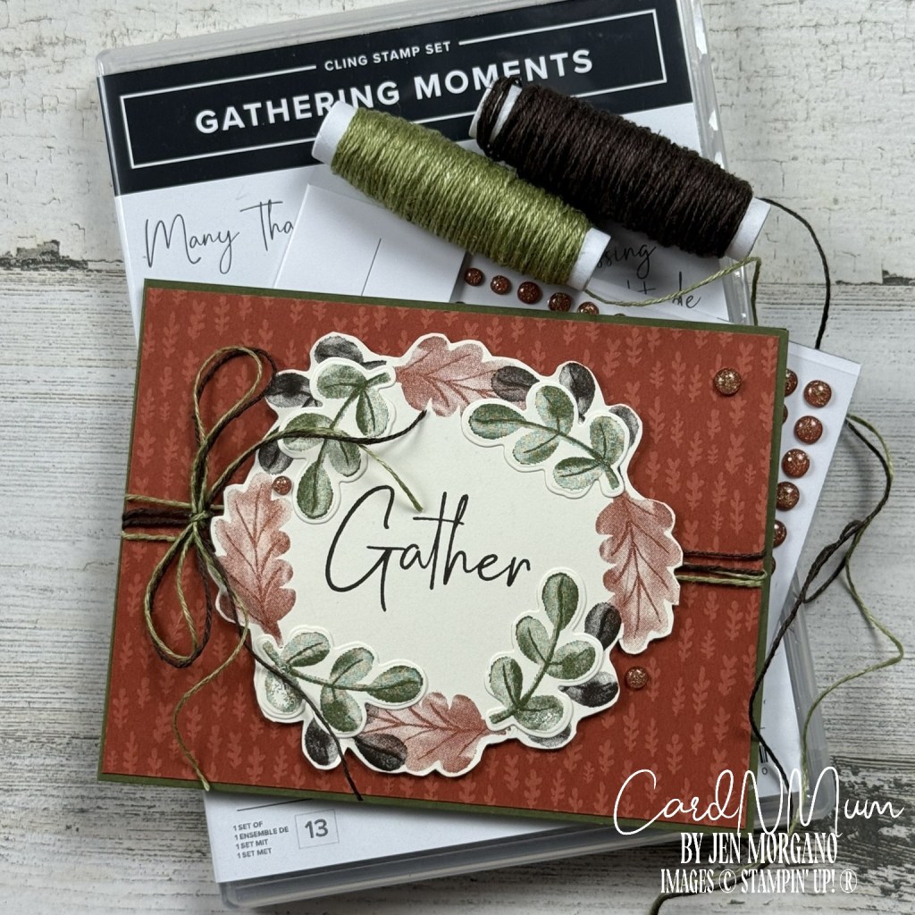 A handmade card featuring a decorative wreath of leaves and the word 'Gather' in the center, placed on top of a patterned card base. The background includes spools of green and brown twine and a stamp set labeled 'Gathering Moments'.