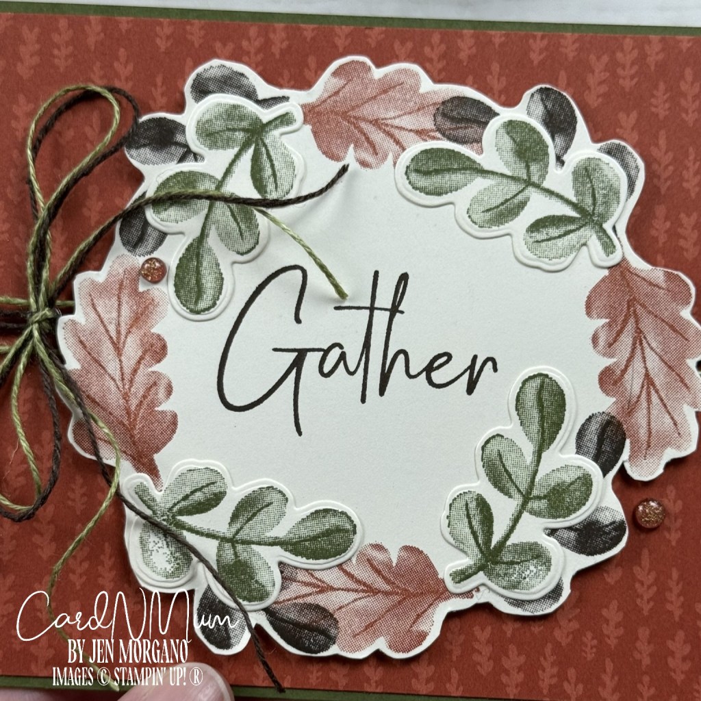 A handmade card featuring a circular wreath design made of various leaf stamps in shades of green, orange, and brown. The center of the card displays the word 'Gather' in a bold font. The background consists of patterned paper in warm tones, with twine wrapped around the design.
