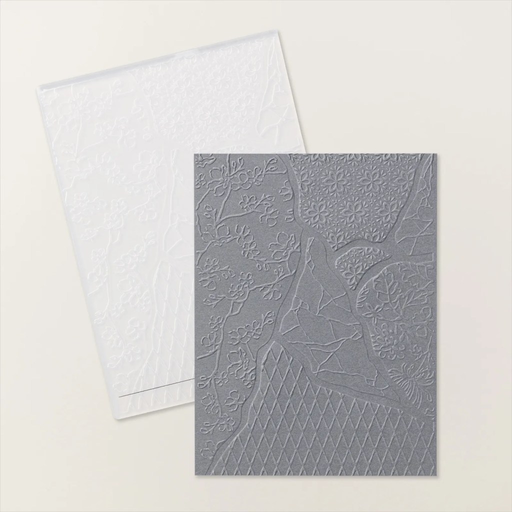 An embossing folder with a detailed floral and geometric design, displayed with a sheet of embossed gray paper.