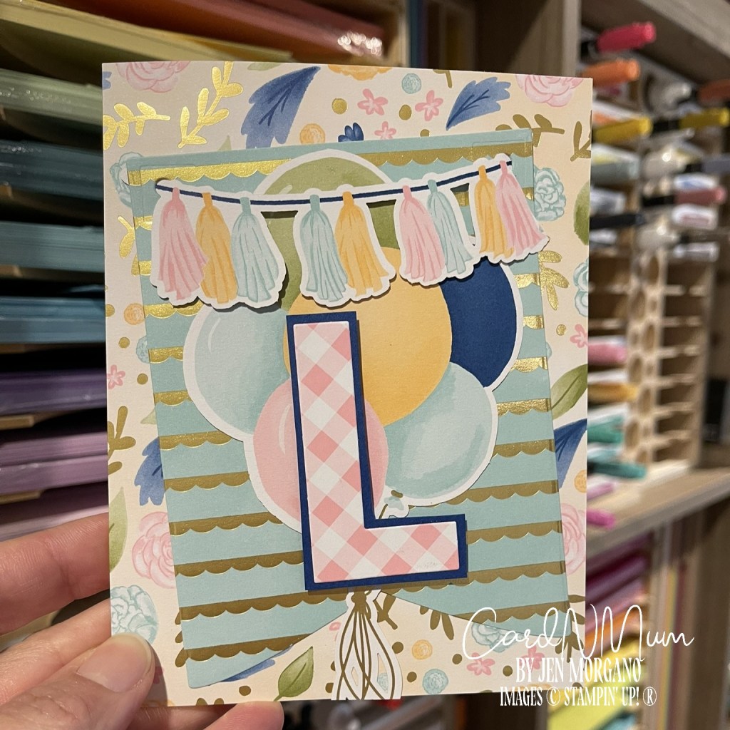 A birthday card featuring the letter 'L' in a plaid design, surrounded by colorful balloons and a decorative banner with tassels. The background has floral patterns and gold accents.