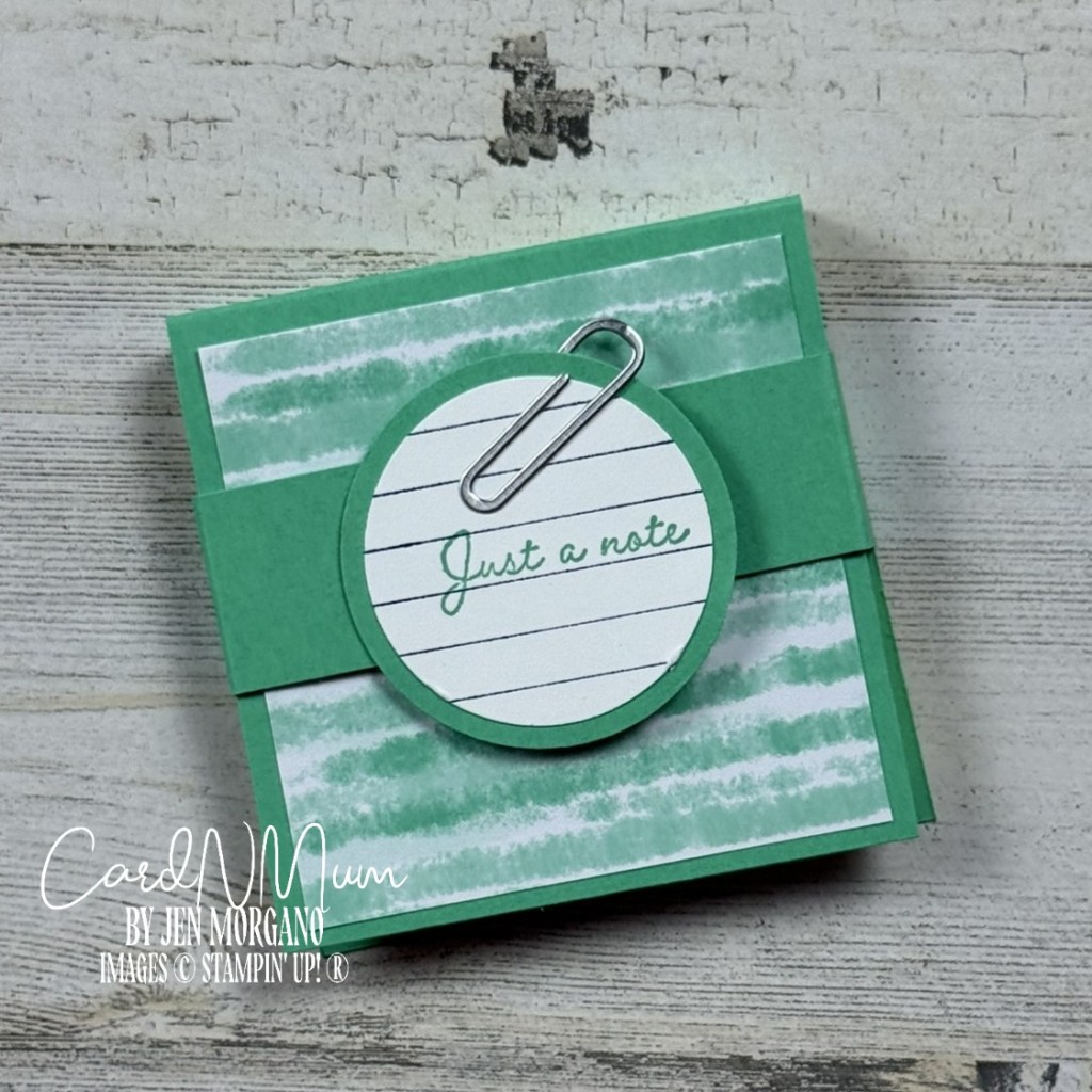 A hand-crafted Post-It Note holder featuring a green and white striped design, with a circular label saying 'Just a note' attached by a silver paperclip.