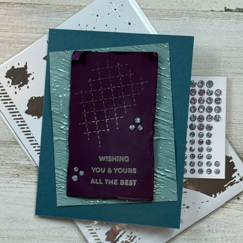 A handmade greeting card featuring a dark purple centerpiece with embossed text 'WISHING YOU & YOURS ALL THE BEST', surrounded by a textured teal border and decorative elements. In the background, there are several sheets of metallic rub-ons and gems.