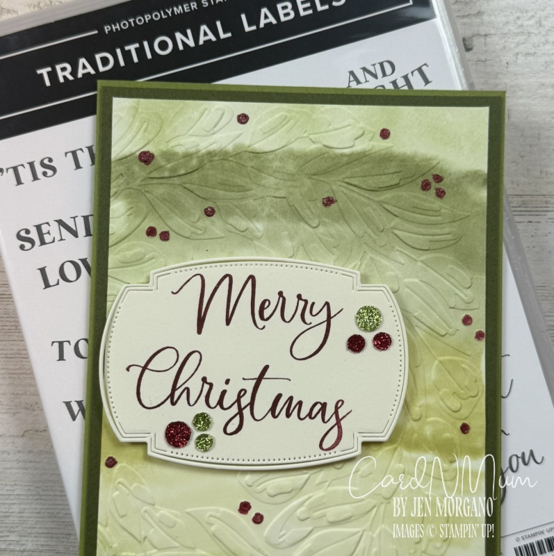 A handmade Christmas card featuring the words 'Merry Christmas' in elegant script, surrounded by green and red glitter embellishments on a textured green background, with a photopolymer stamp set partially visible in the background.