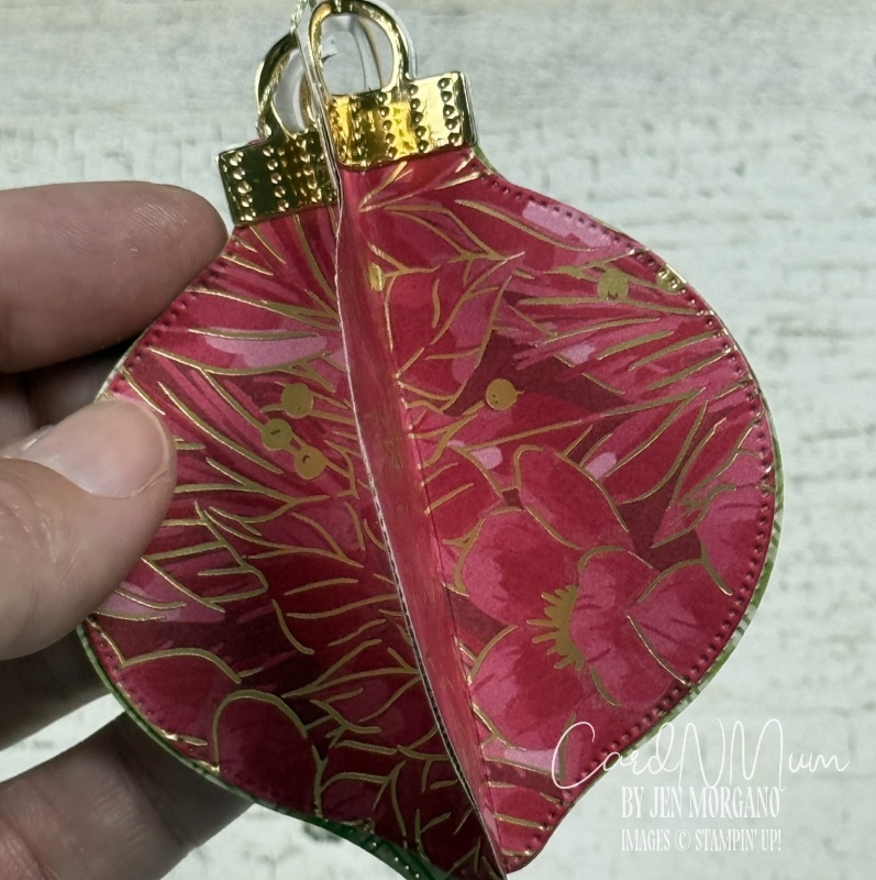A close-up of a decorative leaf-shaped ornament held in a hand, featuring a vibrant pink design with gold accents and floral patterns.