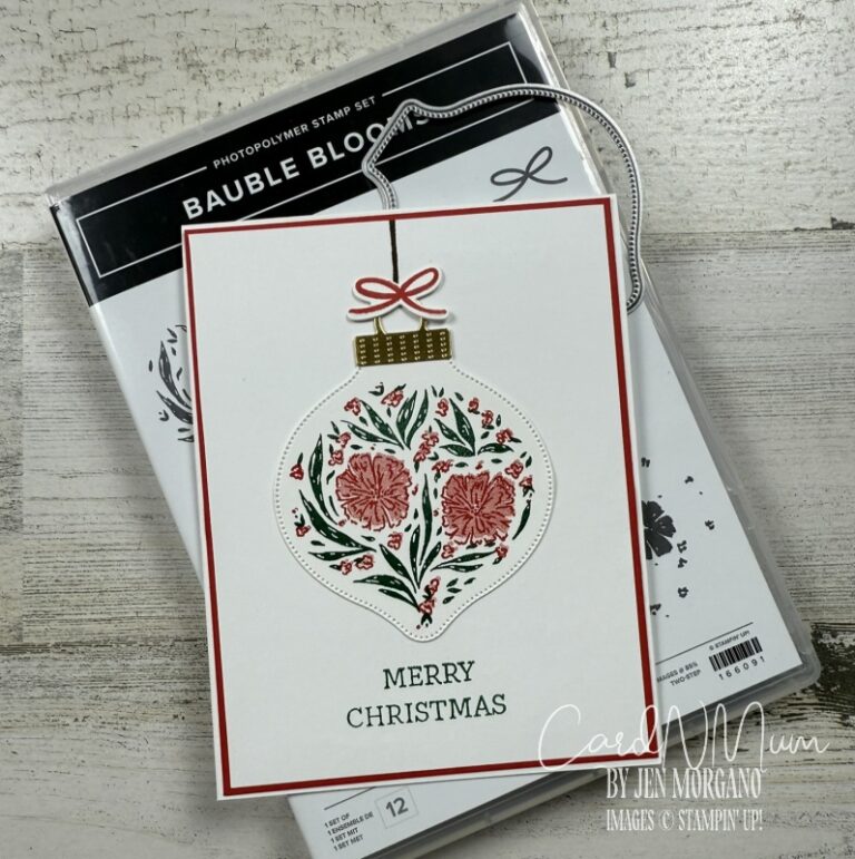 A Christmas card featuring an ornament design with floral elements and the text 'MERRY CHRISTMAS' at the bottom, placed on a wooden surface next to a stamp set package.