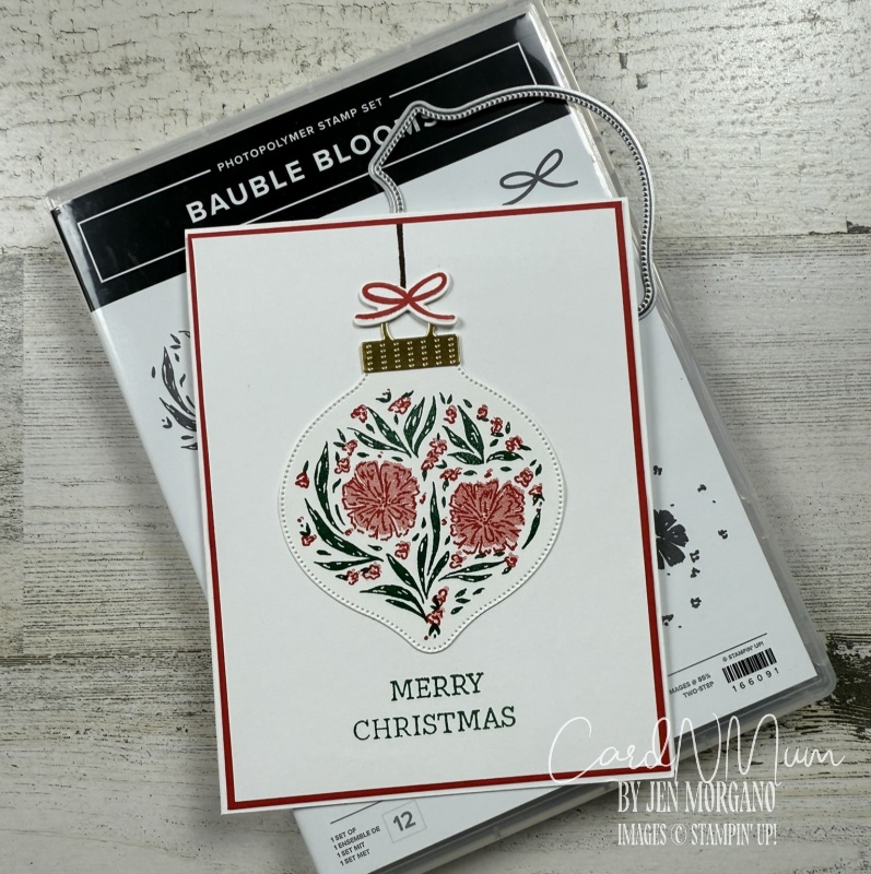 A Christmas card featuring an ornament design with floral elements and the text 'MERRY CHRISTMAS' at the bottom, placed on a wooden surface next to a stamp set package.