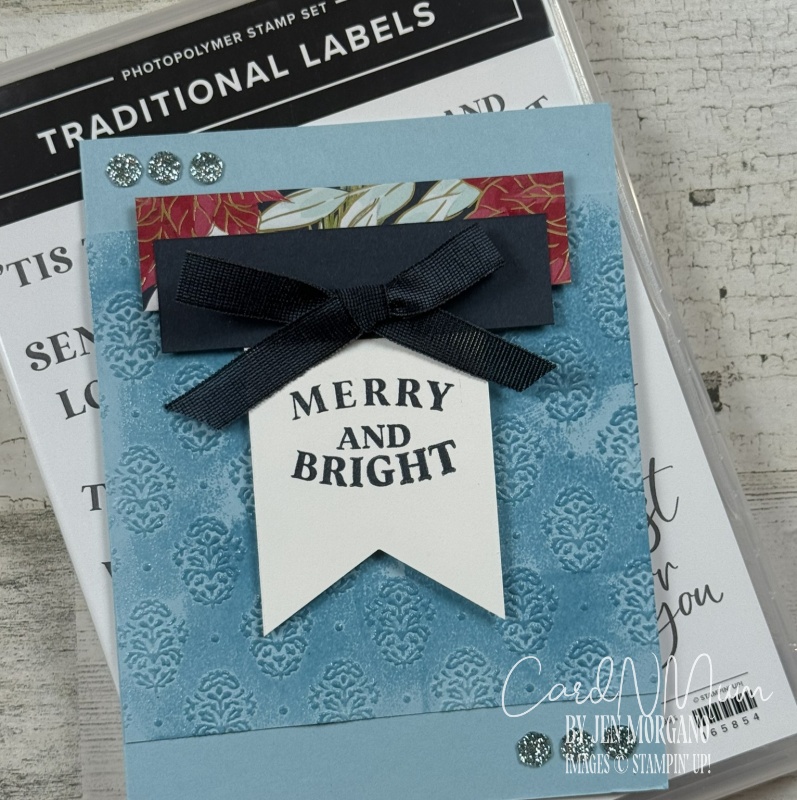 A handmade greeting card featuring a blue embossed background, a dark blue ribbon, and a banner with the text 'MERRY AND BRIGHT'. The card also has decorative elements including patterned paper and glitter accents.